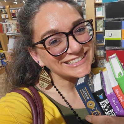 Smiling woman with glasses holding books in a bookstore.
