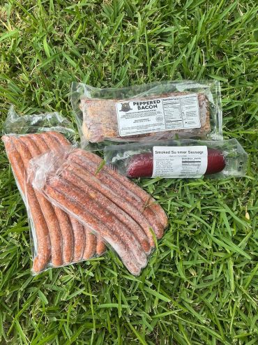 Assorted packaged meats on grass: sausage, peppered bacon, and smoked sausage.