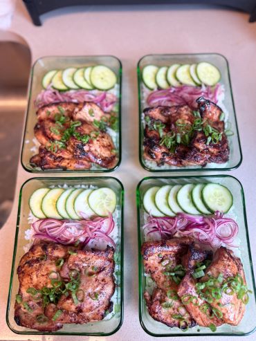 Four meal prep containers with grilled chicken, rice, cucumber, and pickled onions.