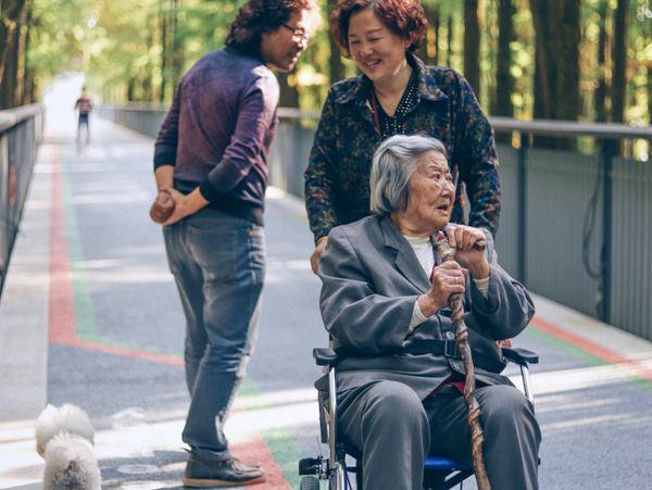 An elderly woman in a wheelchair with two companions and a small dog on a tree-lined path.