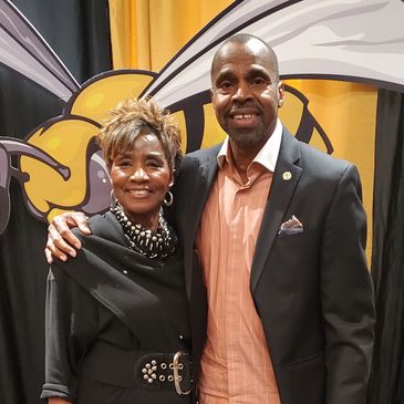 Two people posing in front of a yellow and black backdrop with a bee design.