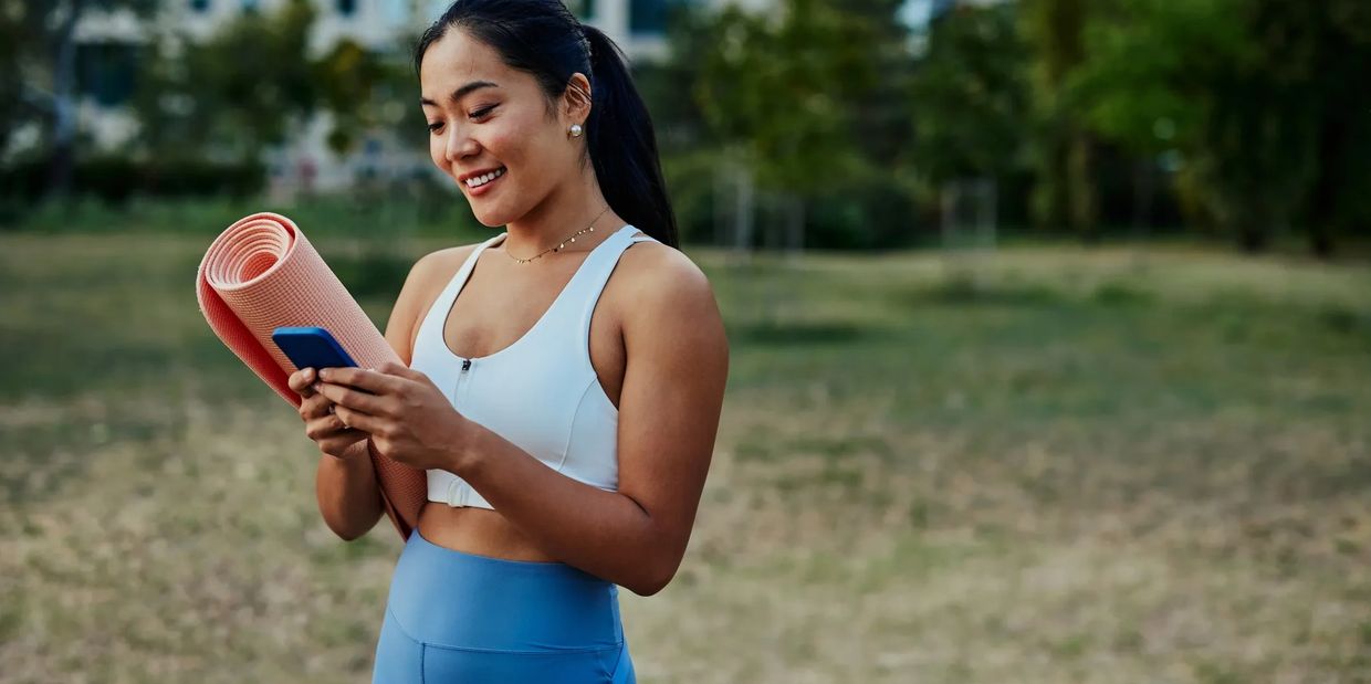 Smiling woman holding a yoga mat and phone outdoors.