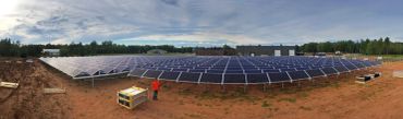 A man stands near solar panels on a large field under a cloudy sky.