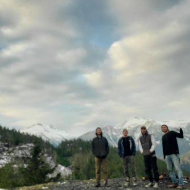 Four people standing outdoors with snowy mountains in the background under a cloudy sky.
