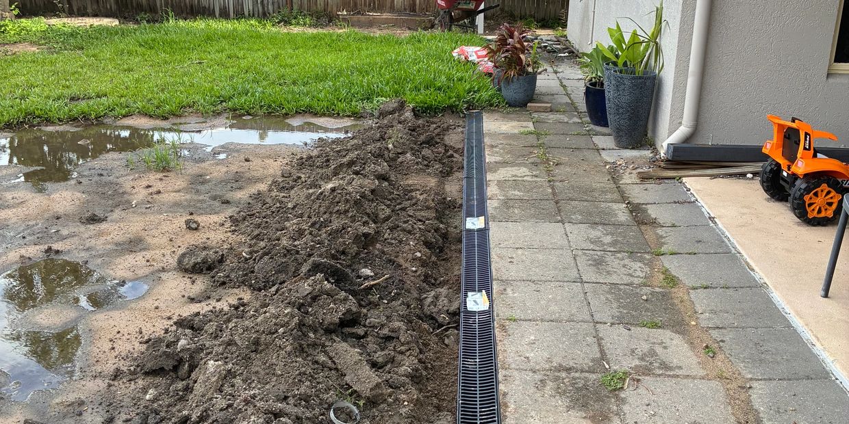 A newly installed drainage grate next to a muddy garden and paved path.