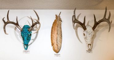 Decorative antler skull sculptures flank a carved wood feather on a gallery wall.