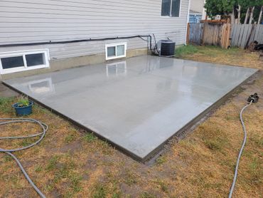 Newly poured smooth concrete patio beside house with garden and fence.