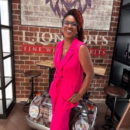 A woman in a bright pink outfit smiles indoors near a vintage car front.