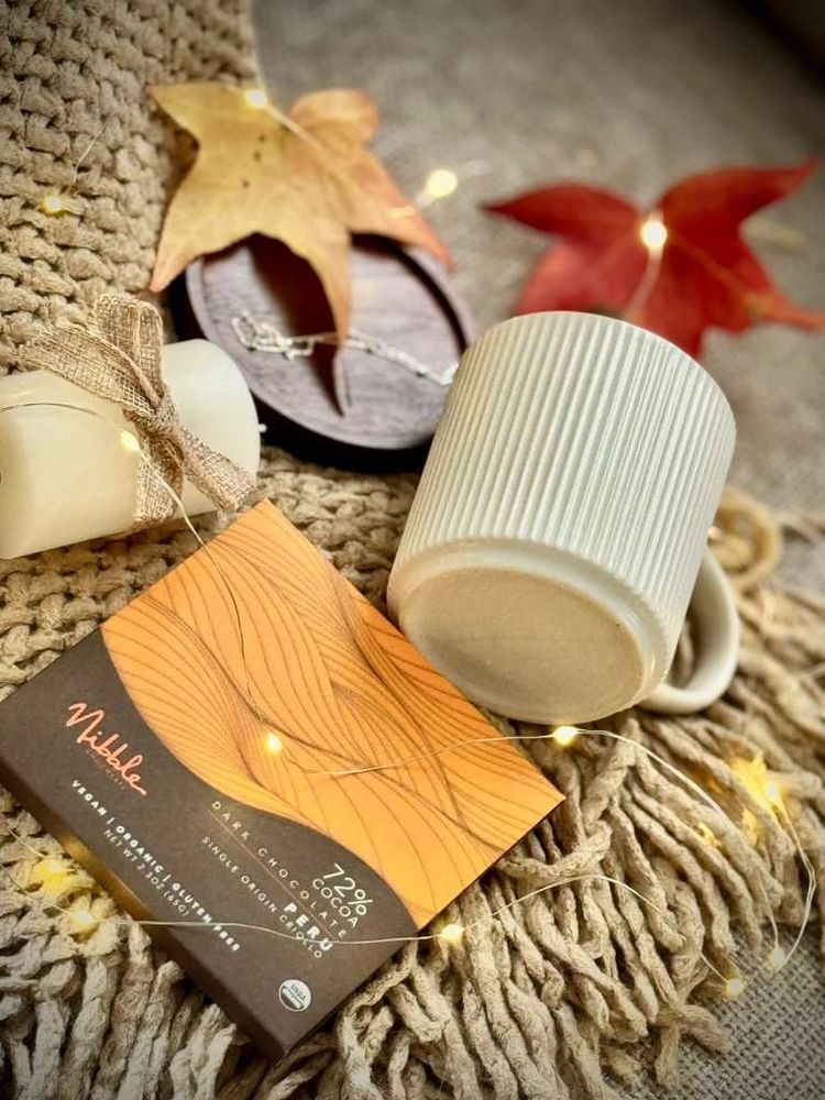 A cozy setup with a ceramic mug, chunky knit blanket, local San Diego vegan chocolate, and a candle.