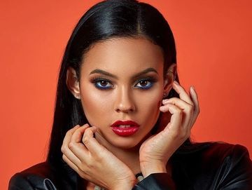 A model poses in a black jacket against an orange backdrop wearing blue eye makeup and red lipstick