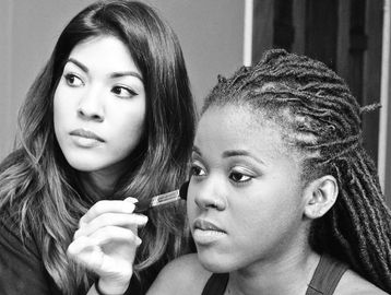 A makeup artist applies makeup to a beautiful dark-skinned model with locs and a nose ring.