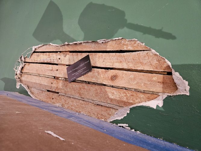 A damaged wall with exposed wood and a business card for drywall repair.