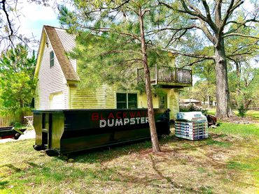 A large Blackwater Dumpster sits beside a yellow house with stacked construction materials.