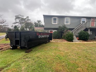 Large black dumpster labeled 'Blackwater Dumpster' near a gray house on a cloudy day.