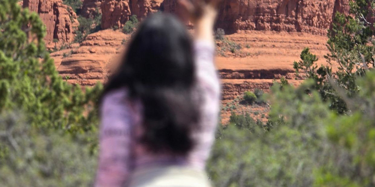 Person with raised hand in front of red rock formations and greenery.