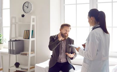 Man consulting doctor, holding neck in pain in a bright medical office.