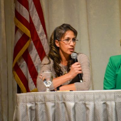 Woman speaking into a microphone at a panel with an American flag behind her.