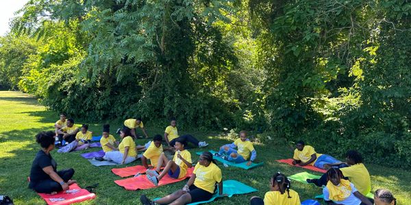 Children in yellow shirts sitting on mats outdoors, listening to an instructor.