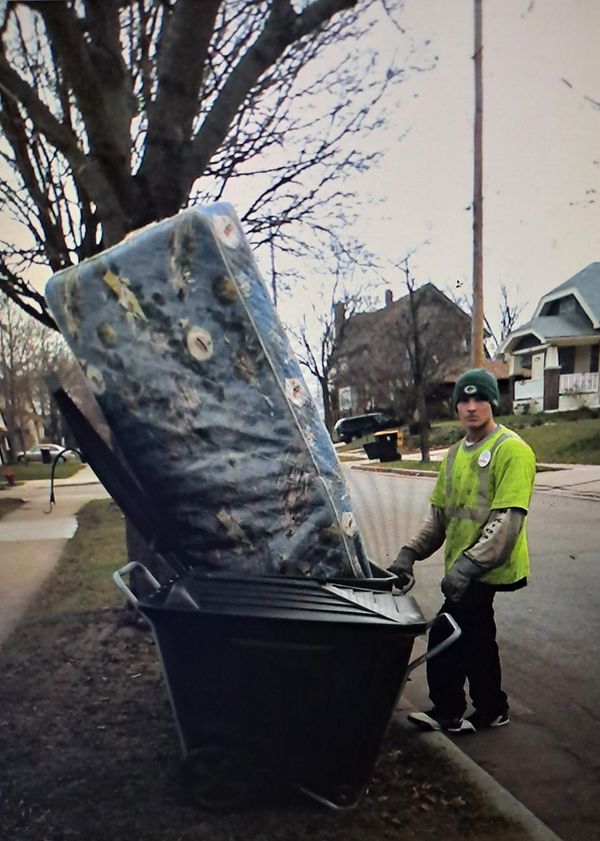Man disposing of a large mattress in a trash bin on a residential street.
