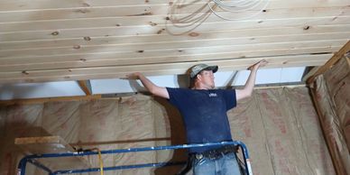 A man installs wooden paneling on a ceiling inside a house under construction.