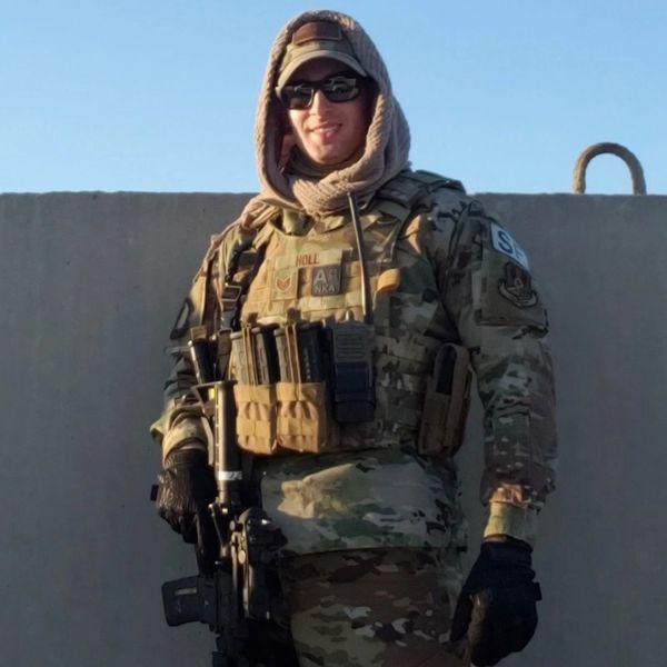 Soldier in camouflage gear holding a rifle and smiling.