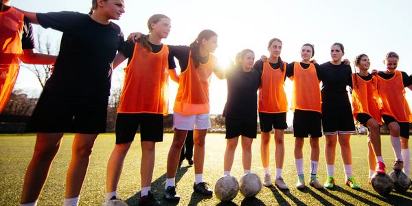A girls' soccer team stands in a circle with arms around each other on a sunny field.