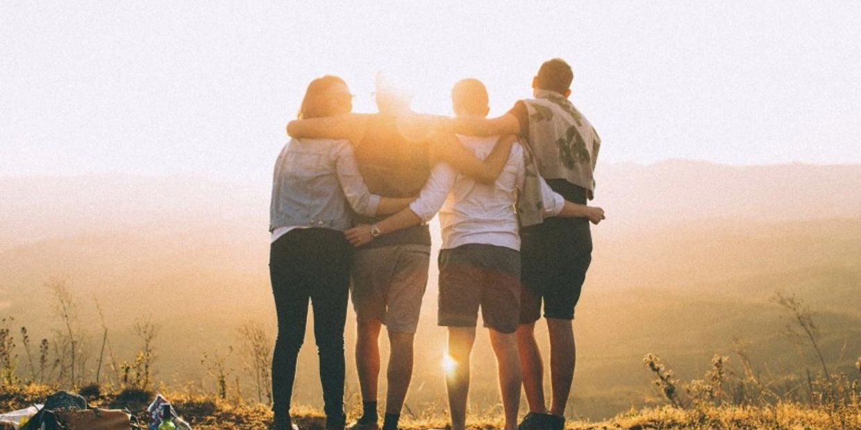 Four friends embracing each other, watching the sunset over a scenic landscape.