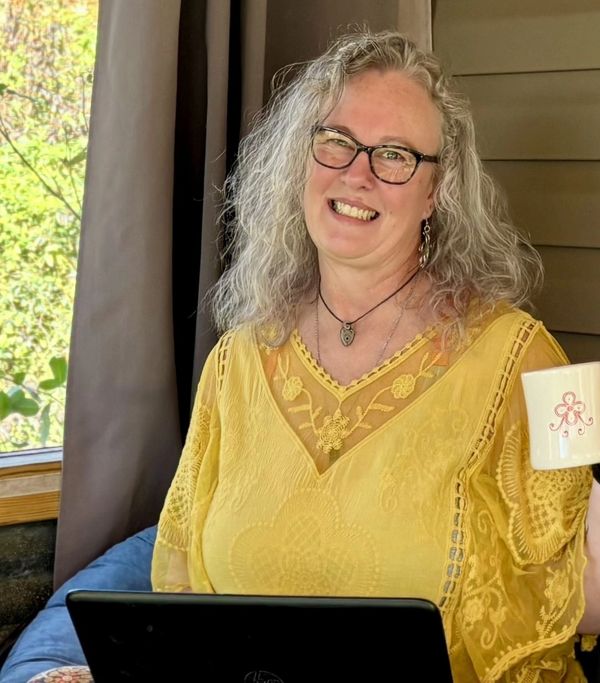Smiling woman in glasses and a yellow blouse holding a coffee mug with a laptop.