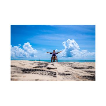 Man in wheelchair on a sunny beach with arms outstretched.