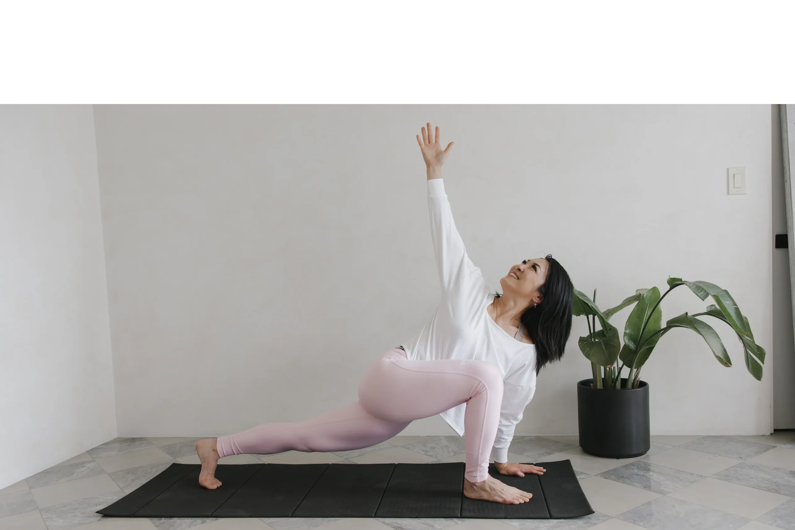 J.J. stands on a yoga mat with a plant behind her. She is in a lunge and reaches her hand upward.