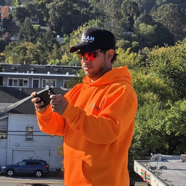 Man in orange hoodie and black cap using a smartphone outdoors.