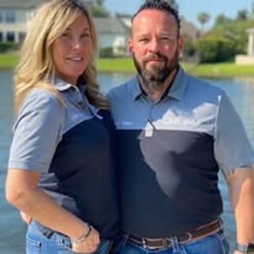 A smiling couple stands outdoors near water, both wearing matching polo shirts.