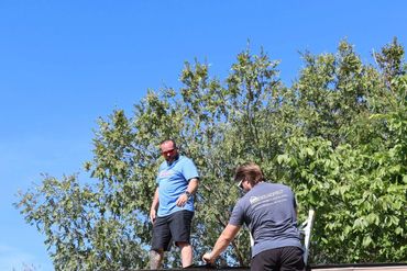 Two men working on a roof under a clear blue sky, one on ladder and one standing on roof.