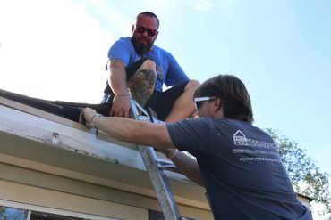 Two men working on a roof using a ladder under clear sky.