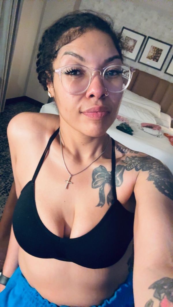Young woman with glasses and tattoos takes a selfie in a bedroom.