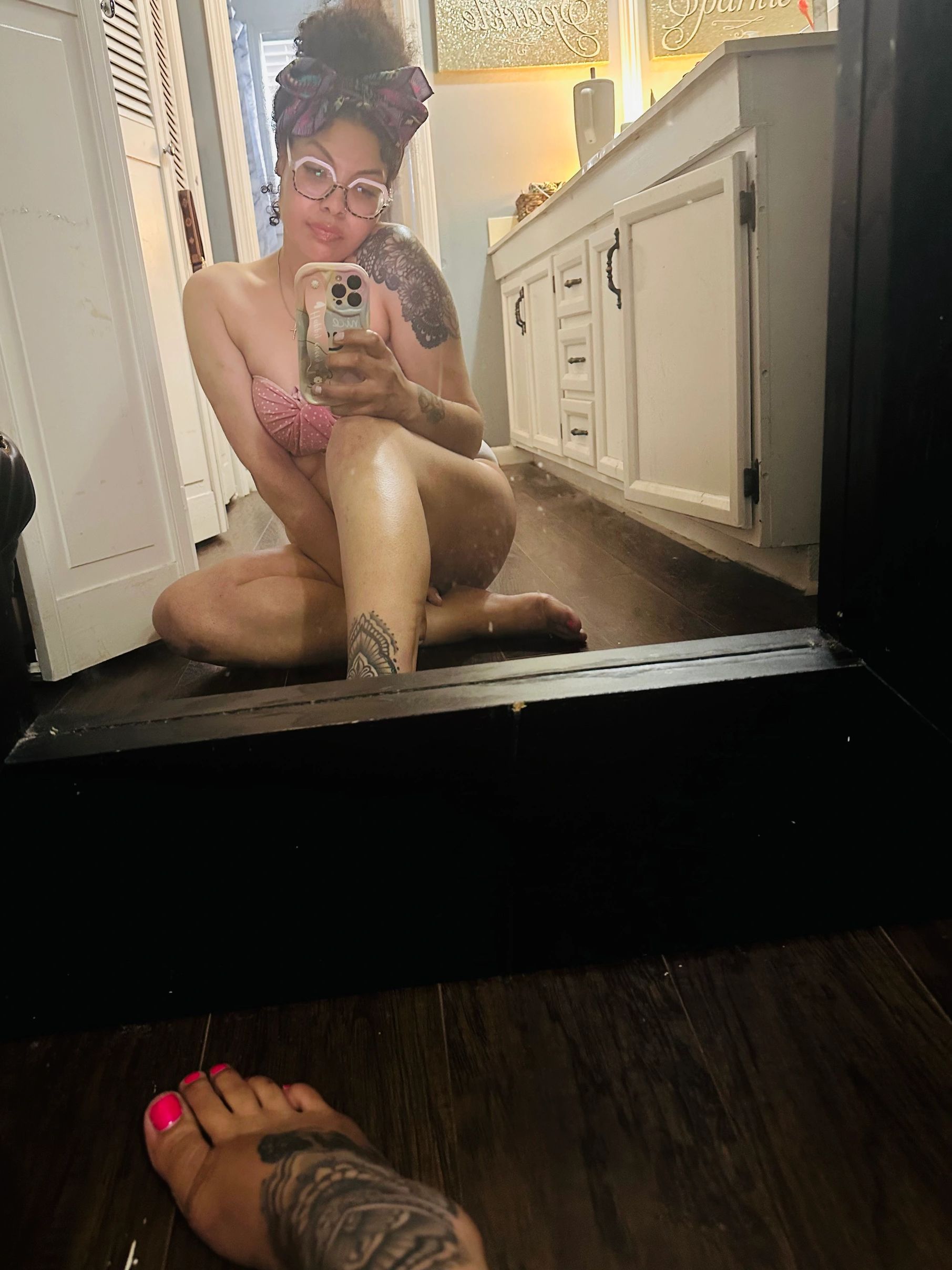 Woman in glasses takes mirror selfie sitting on the floor with tattoos and pink nails.
