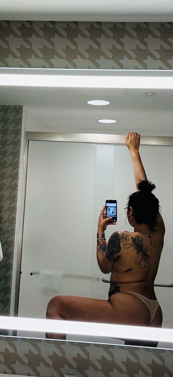 A tattooed woman takes a mirror selfie in a bathroom wearing minimal clothing.