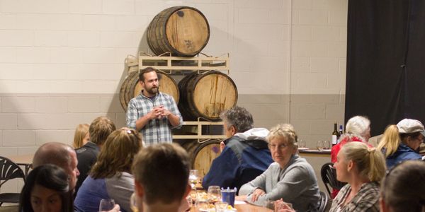 Man speaking to a group at a winery with barrels in the background.