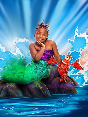 A girl dressed as a mermaid with a crown and a cartoon crab on a rock.