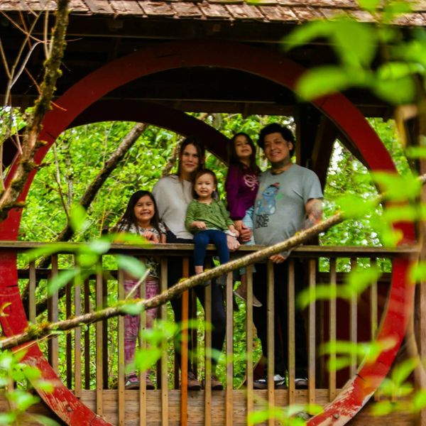 A family of five stands on a wooden balcony framed by a circular red arch surrounded by greenery.