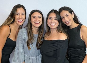 Four women smiling and posing together against a plain background.