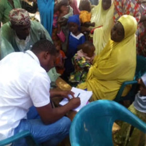 A man in white registers or interviews a woman in yellow surrounded by children and adults.