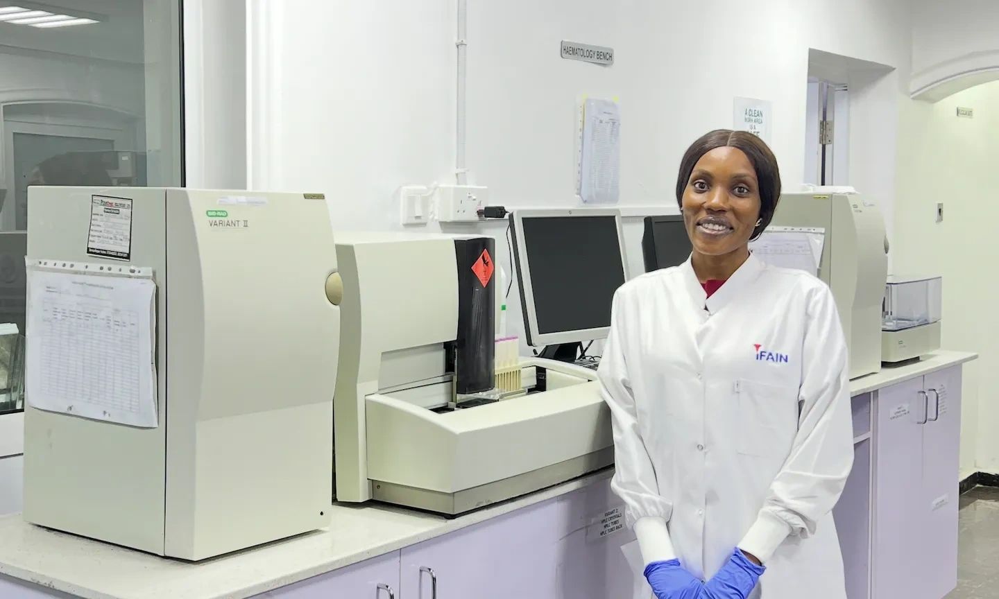 A smiling lab technician in a white coat and blue gloves stands in a modern laboratory.