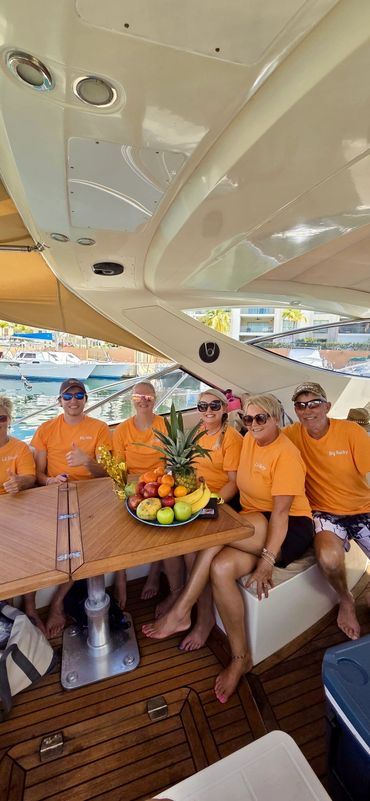 Group of six people in matching orange shirts sitting on a boat with a fruit plate.