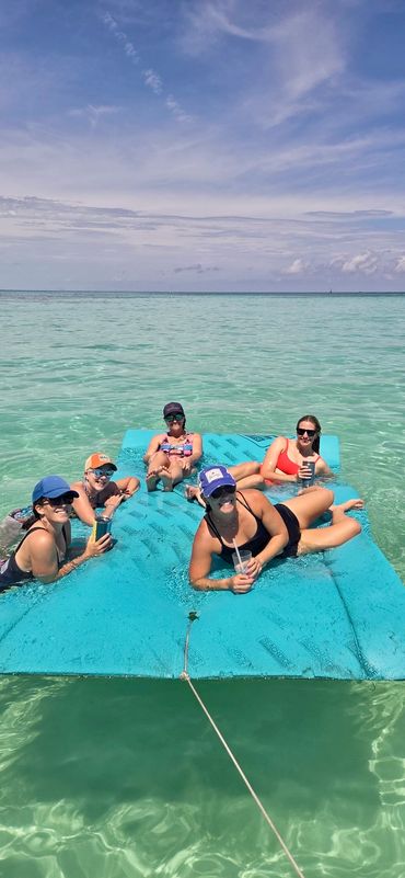 Five women relaxing on a floating mat in turquoise sea water under a blue sky.