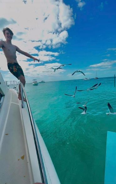A young man on a boat enjoying a sunny day with seagulls flying over turquoise water.