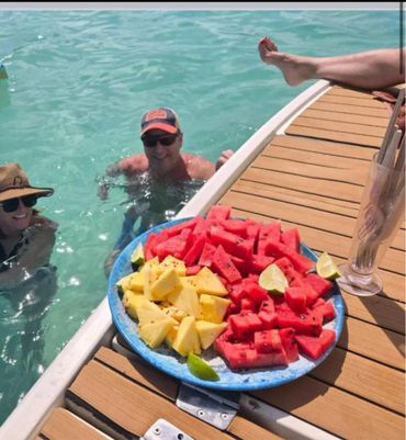 People enjoying swimming near a dock with a plate of fresh pineapple and watermelon.