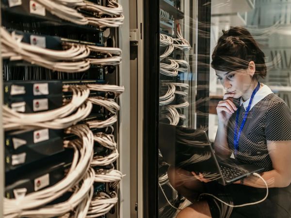Woman working on a laptop inside a server room with network cables.