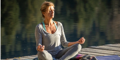 A woman relaxes in meditatation on a dock by a lake surrounded by trees