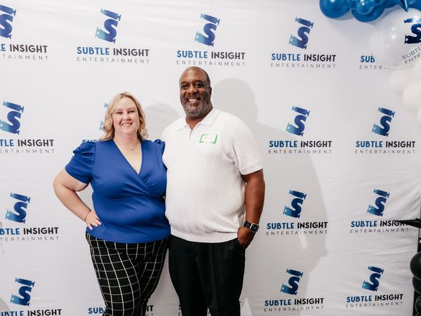 Two smiling people posing in front of a Subtle Insight Entertainment backdrop.